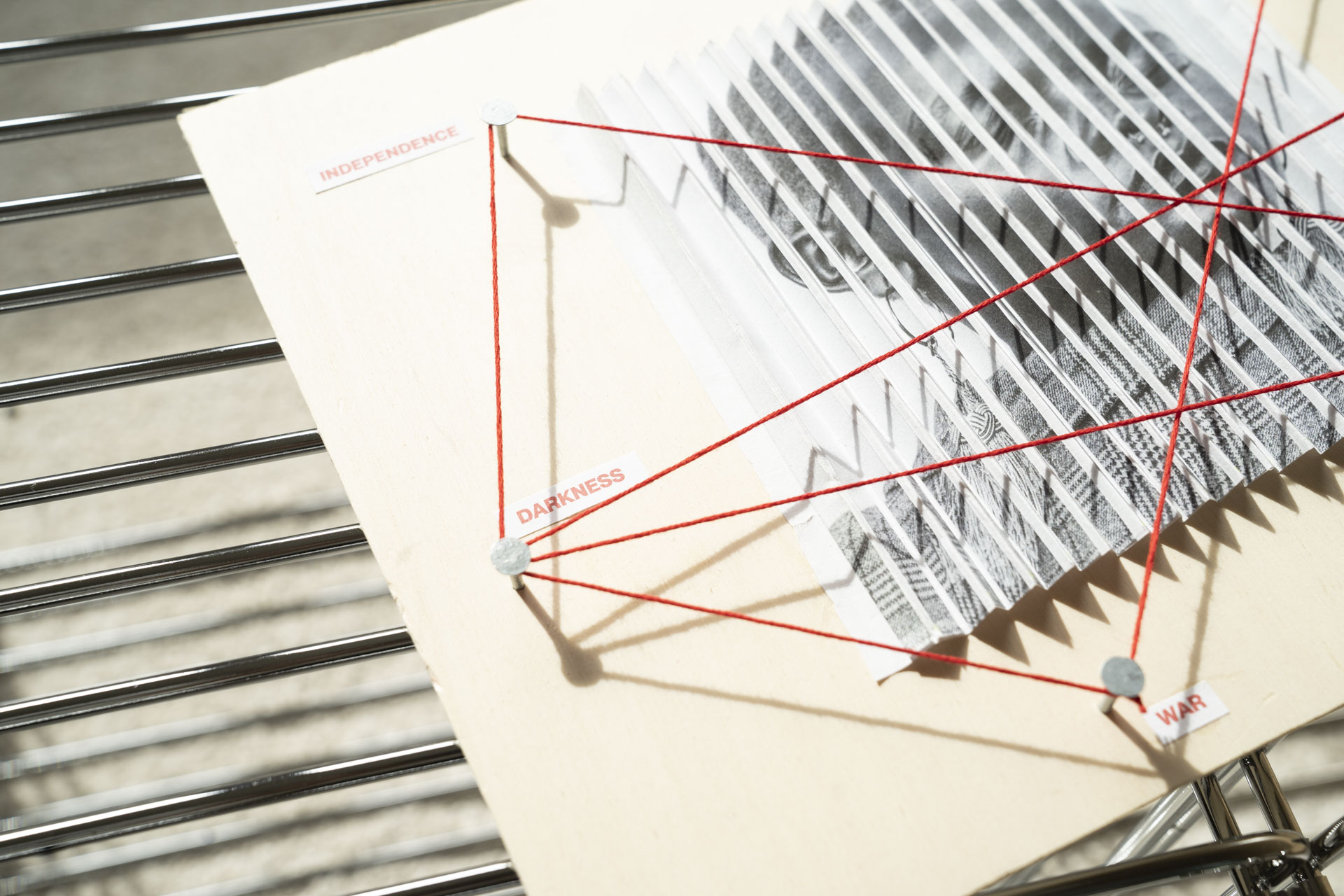 Detailaufnahme eines Holzbretts mit einem gefalteten Blatt Papier mit einem Bild darauf. Über dem Bild ein roter Faden mittels Nägel gespant, welcher mehrere Begriffe zu alltagsrelevanten Themen verbindet.