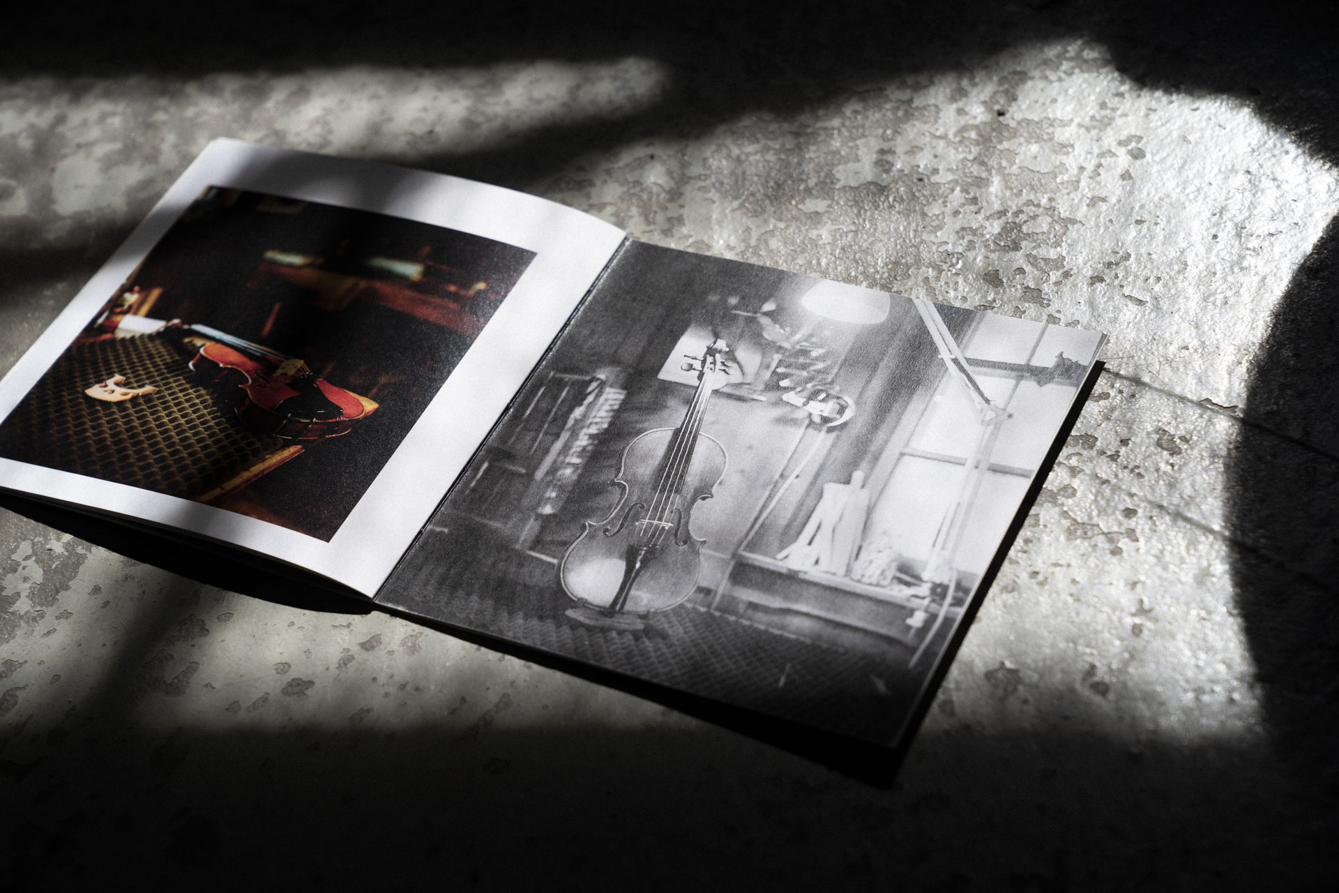 Geöffnetes Violin Maker Fotobuch auf einem rustikalen Steinboden mit Schatten. Ein Foto einer Violine ist abgebildet.