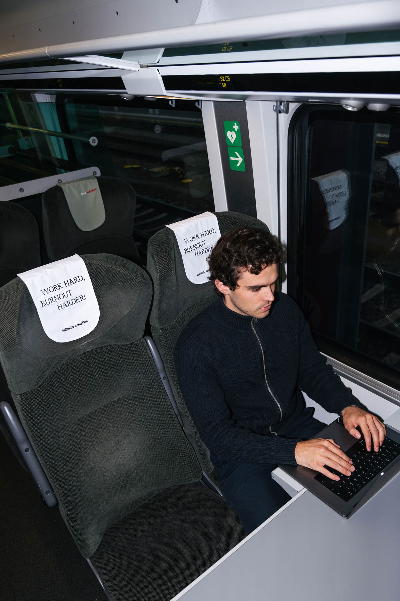 Sitzplatz mit Tisch in einem Railjet. Person sitzt mit Laptop und arbeitet. Das Kopfteil des Sitzes zeigt die Aussage Work Hard, Burnout Harder