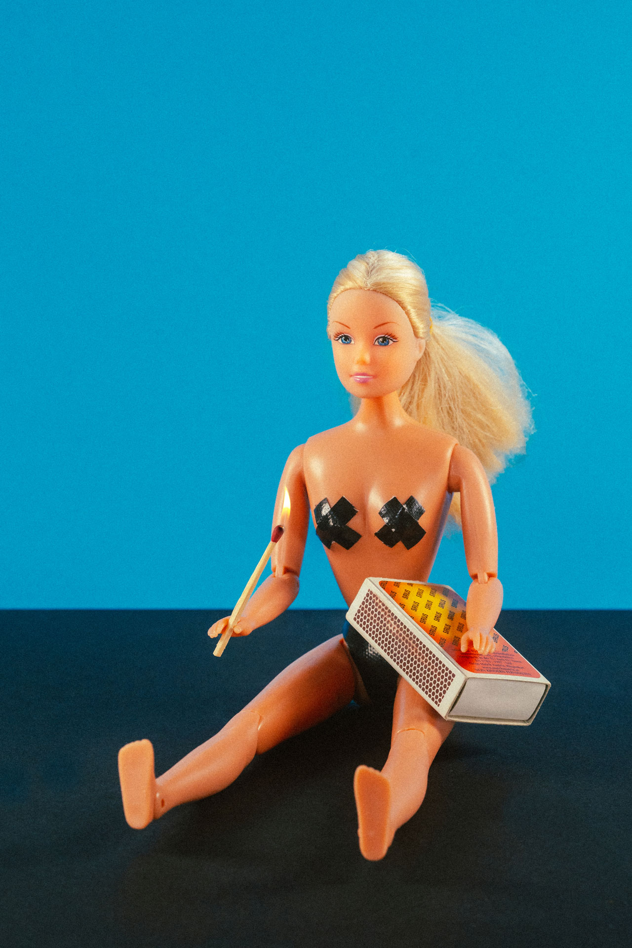 Nahaufnahme einer modifizierten Barbie mit einer Streichholzschachtel und brennenden Streichholz in der Hand